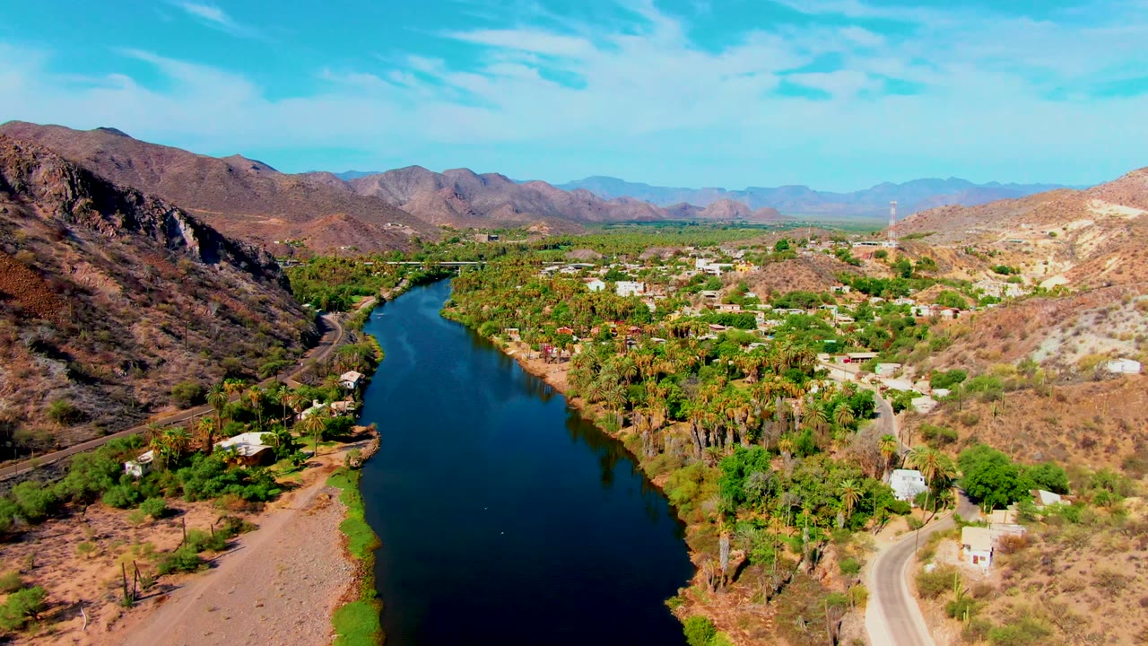 Mulege aerial view with the river running up the left side, a rocky brown hillside on the right, green trees around homes in the center, distant mountains, and no visible people.