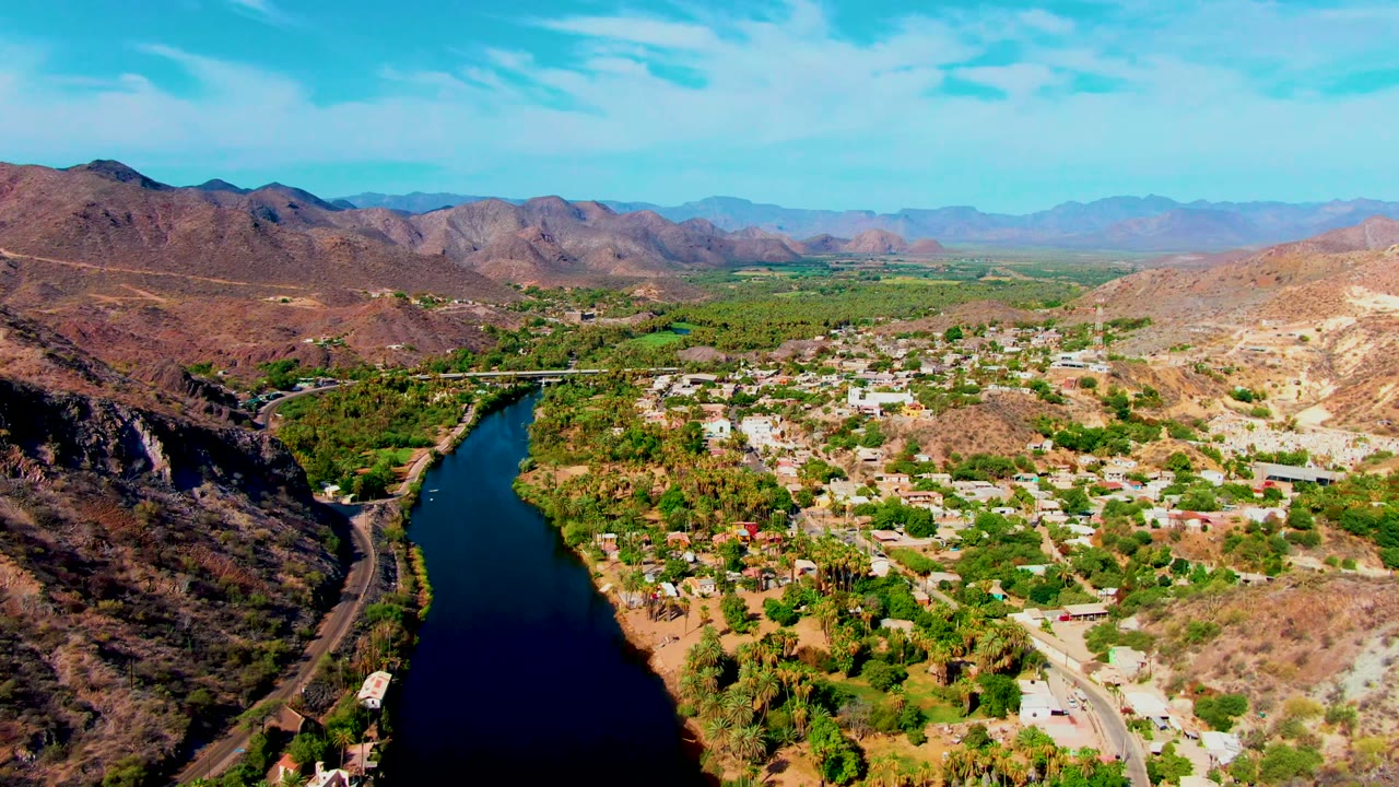 Opening aerial frame over Mulege, Baja California Sur, showing a dark river on the left, green riverbank trees, a small town, brown hills, distant mountains, blue sky, and no visible people.