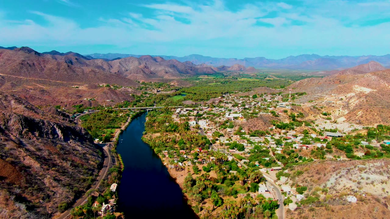 Opening aerial frame over Mulege, Baja California Sur, showing a dark river at left, green trees around scattered buildings, dry hills, distant mountains, blue sky, and no visible people.