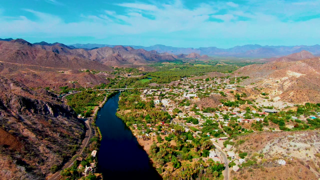 Wide drone view of a river, road, green trees, and dry hills around a town.