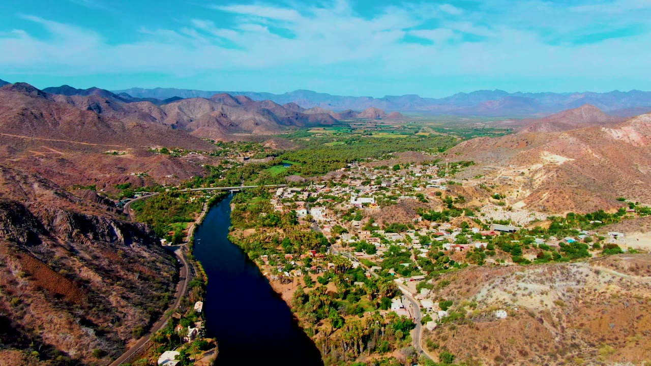 Aerial view over Mulege, Baja California Sur, with a dark river at lower left, green riverbank trees, scattered buildings, dry hills, distant mountains, and no visible people.