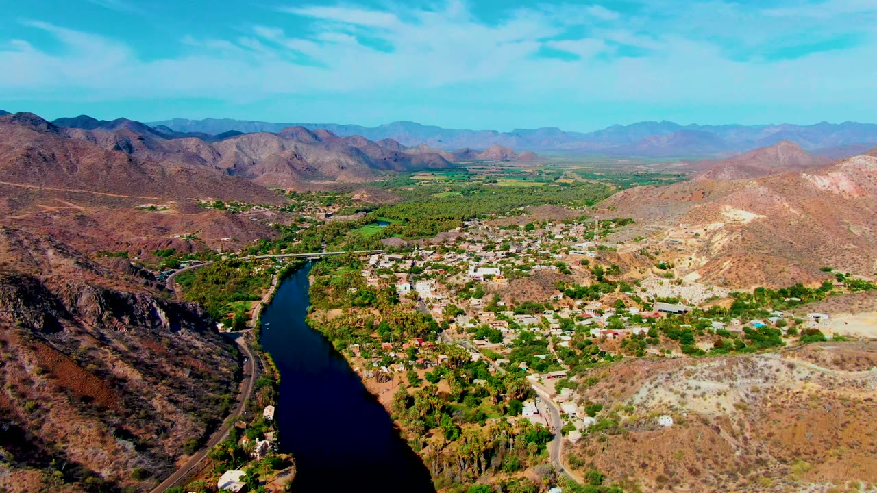 Wide aerial frame in Mulege showing the river curving below dark hills, dense green vegetation along the water, pale town buildings, dry slopes, and no visible people.