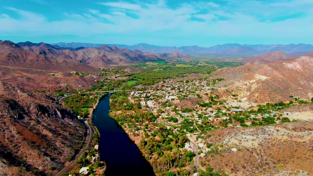 Aerial view across Mulege with the river and bridge toward the left, a spread of low buildings and trees in the center, tan hills to the right, distant ridges, and no visible people.