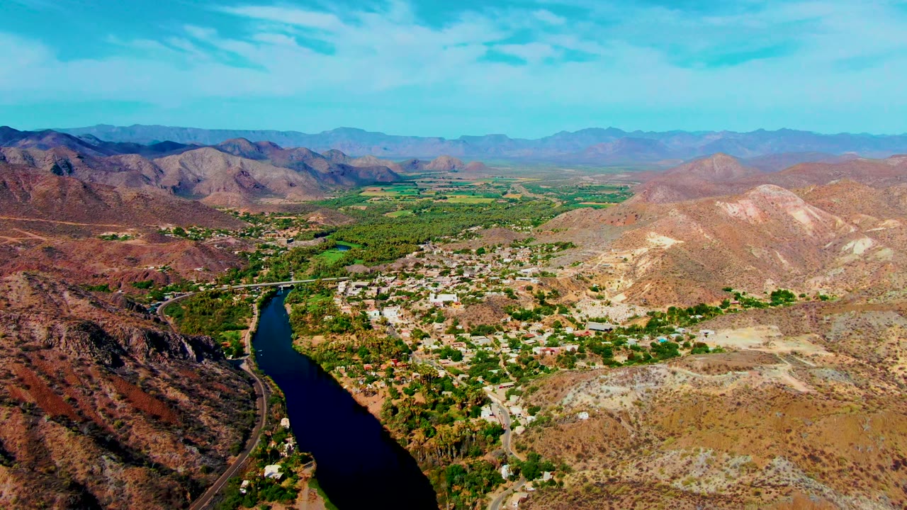 Wide aerial over river, settlement, green valley, and mountains