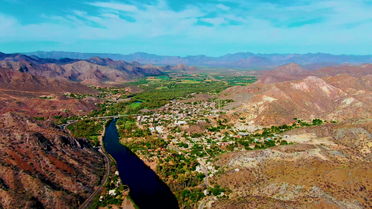Aerial landscape over Mulege with the dark river running up from the lower left, a compact town area beside green vegetation, pale brown hills in the center, distant mountains, and no visible people.