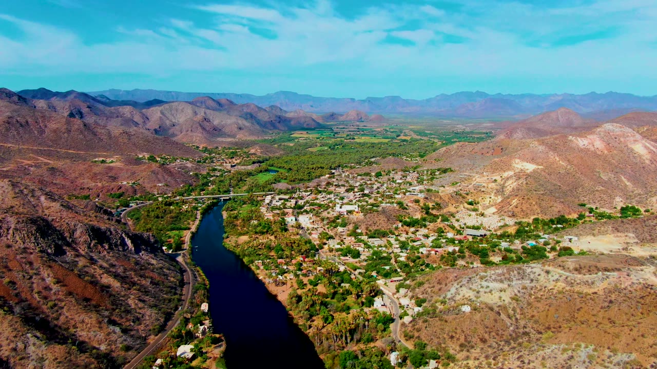 Opening aerial frame over Mulege, Baja California Sur, with a dark river at left, a bridge near the settlement, arid hills, distant mountains, blue sky, and no visible people.