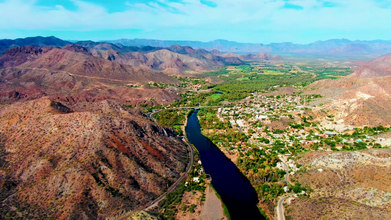 Opening aerial frame over Mulege, Baja California Sur, showing a dark river at lower left, pale town buildings, green vegetation, dry hills, distant mountains, and no visible people.