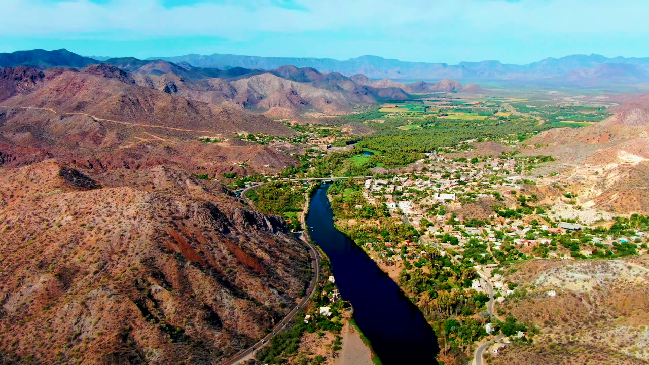 Mulege appears from above with a bridge crossing the river near the lower center, green fields and trees spreading into the valley, reddish hills, far mountains, and no visible people.
