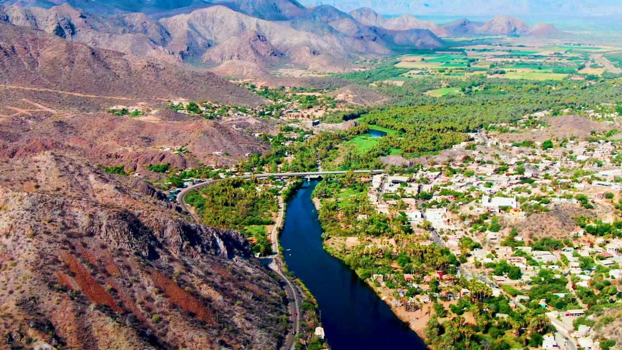 High aerial view over Mulege, Baja California Sur, showing a dark river curving past a town, bridge, green trees, dry hills, and distant mountains, with no visible people.