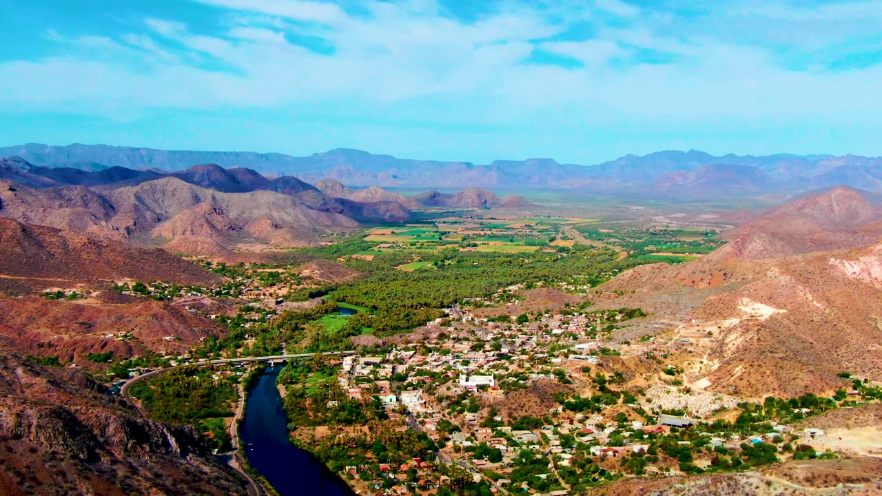 Opening aerial frame over Mulege, Baja California Sur, with a dark river curving below a small town, arid hills, distant mountains, blue sky, and no visible people.