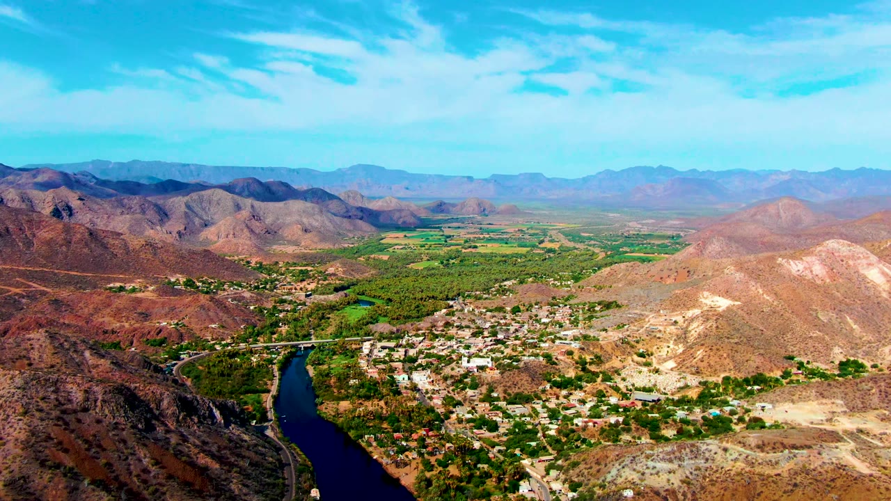 Third aerial frame over Mulege, Baja California Sur, with pale hills in the foreground, a dark river bend at lower right, dense green patches along the valley, distant mountains, and no visible people.