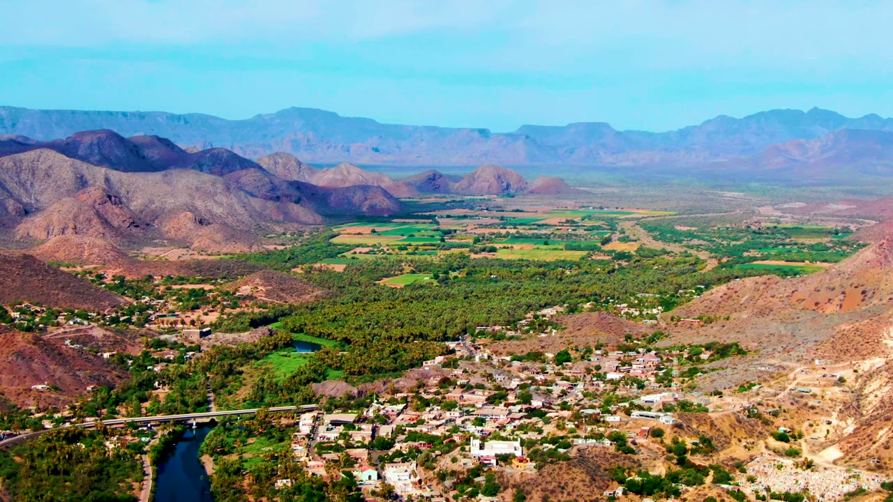 Opening aerial frame over Mulege, Baja California Sur, Mexico with a river bend at lower left, clustered light buildings, green valley fields, brown hills, distant mountains, blue sky, and no visible people.
