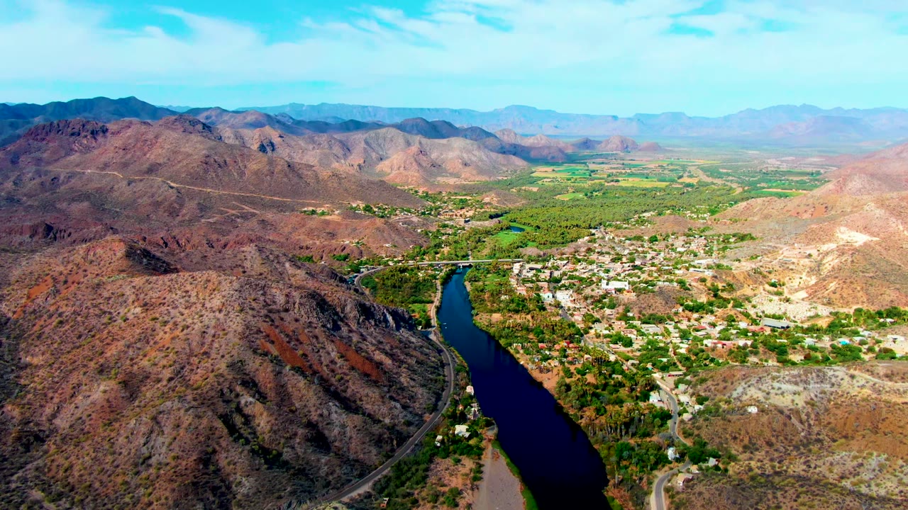 High aerial view over Mulege, Baja California Sur, with a dark river below, a winding road on the hillside, a small town, green valley fields, rocky mountains, bright sky, and no visible people.