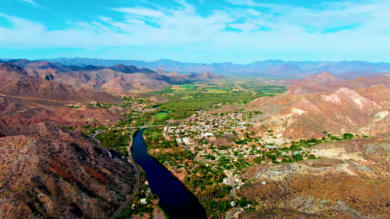 Wide aerial view over Mulege, Baja California Sur, with a dark river at lower left, green valley trees, pale hillside settlement, dry mountains, blue sky, and no visible people.