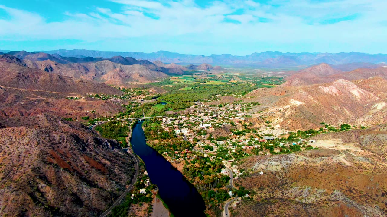 Aerial view across Mulege, Baja California Sur, showing a narrow river below, dense green vegetation along the valley floor, pale buildings, tan ridges, hazy mountain layers, and no visible people.