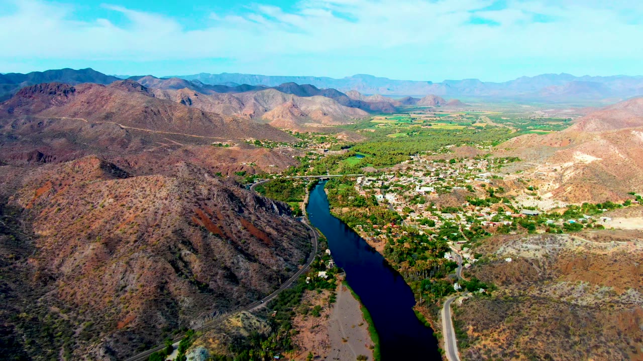 Opening aerial frame over Mulege, Baja California Sur, showing a dark blue river between rocky hills, green riverside vegetation, distant settlement buildings, and no visible people.