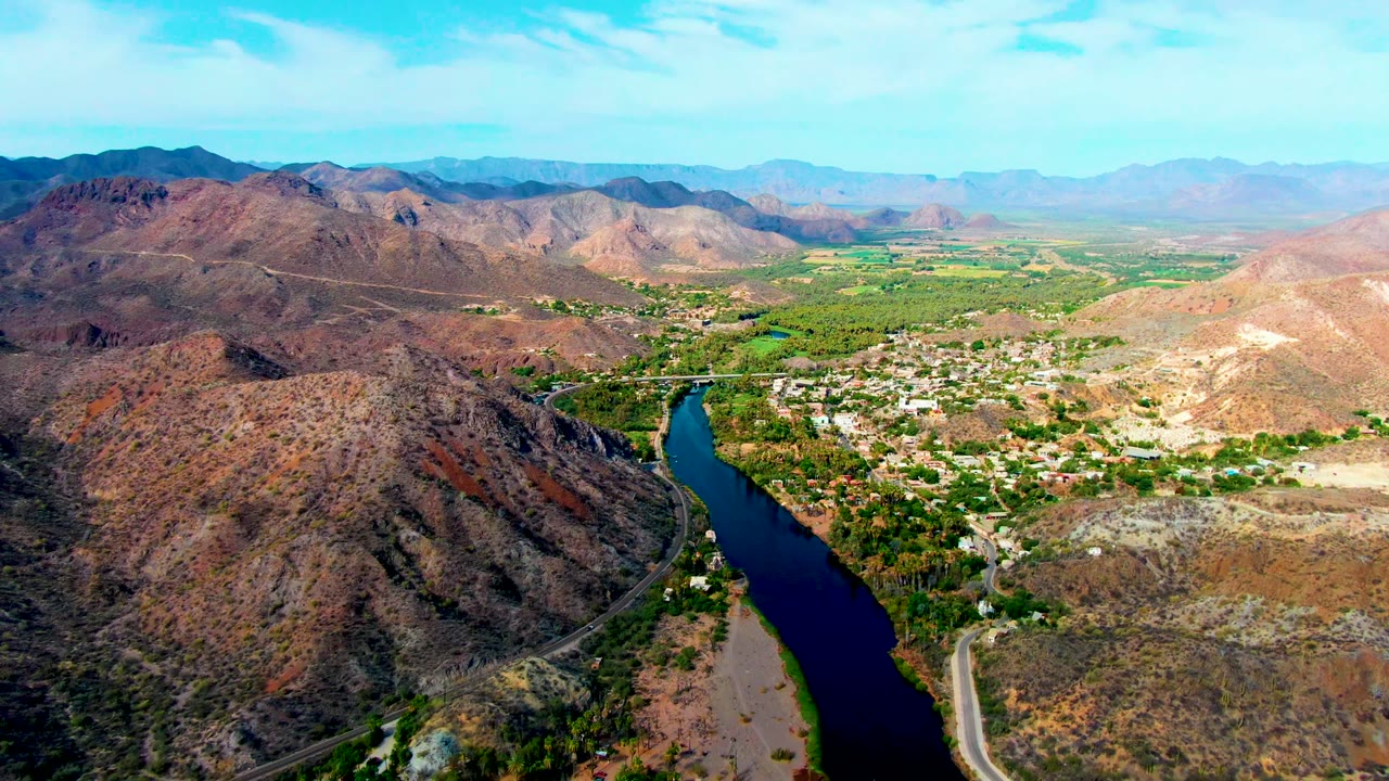Drone view of a blue river, rocky hills, roads, and a green valley settlement.