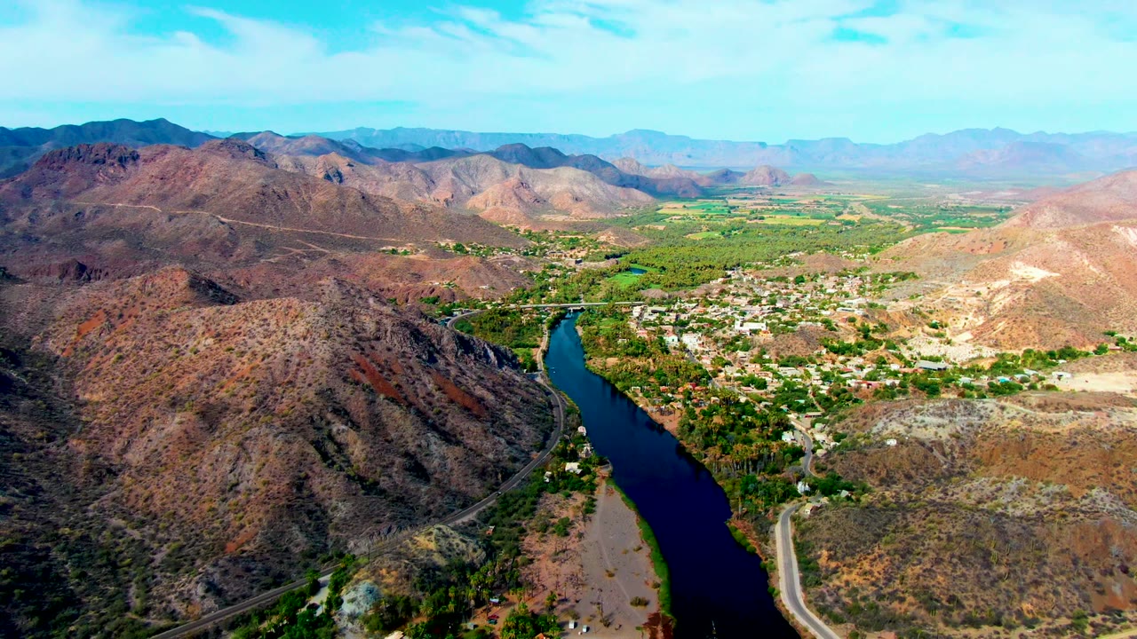 Wide Mulege aerial frame showing the river bend at lower center, green valley vegetation, pale town blocks, reddish brown hills, and hazy mountain ridges with no visible people.