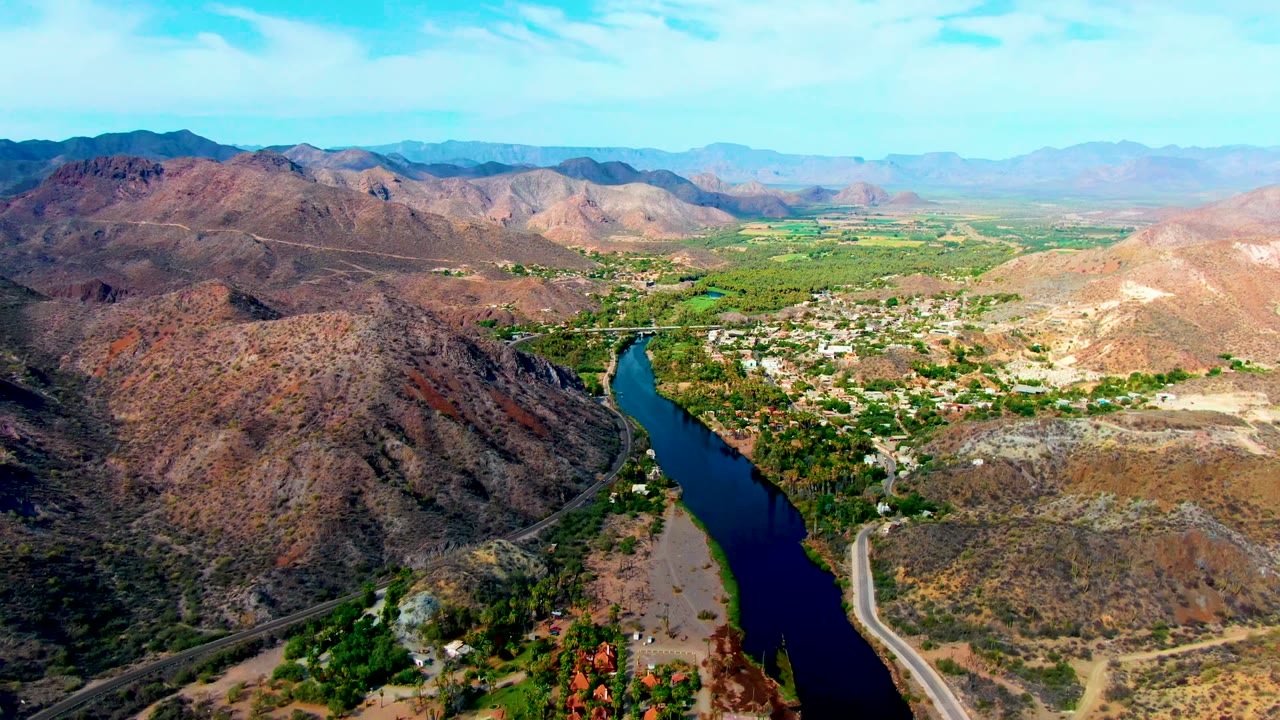 Opening aerial frame over Mulege, Baja California Sur, showing a dark river curving between brown hills, green trees, pale roads, distant mountains, and no visible people.