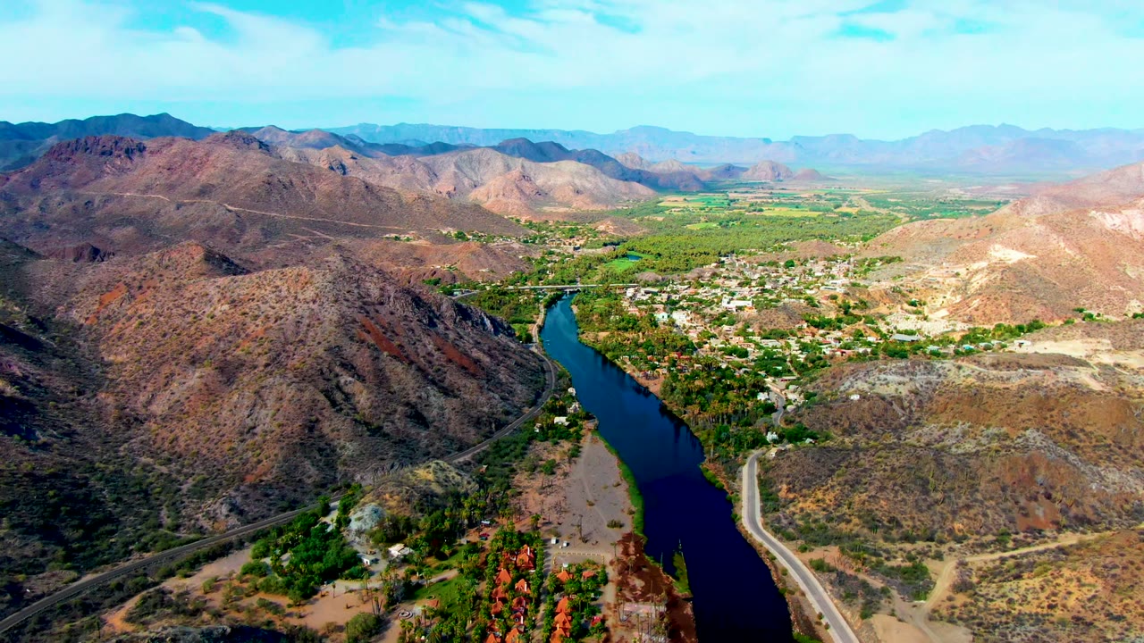 Wide aerial frame over Mulege, Baja California Sur, showing the blue river below a rocky hillside, green fields and trees around a small settlement, distant mountains, and no visible people.