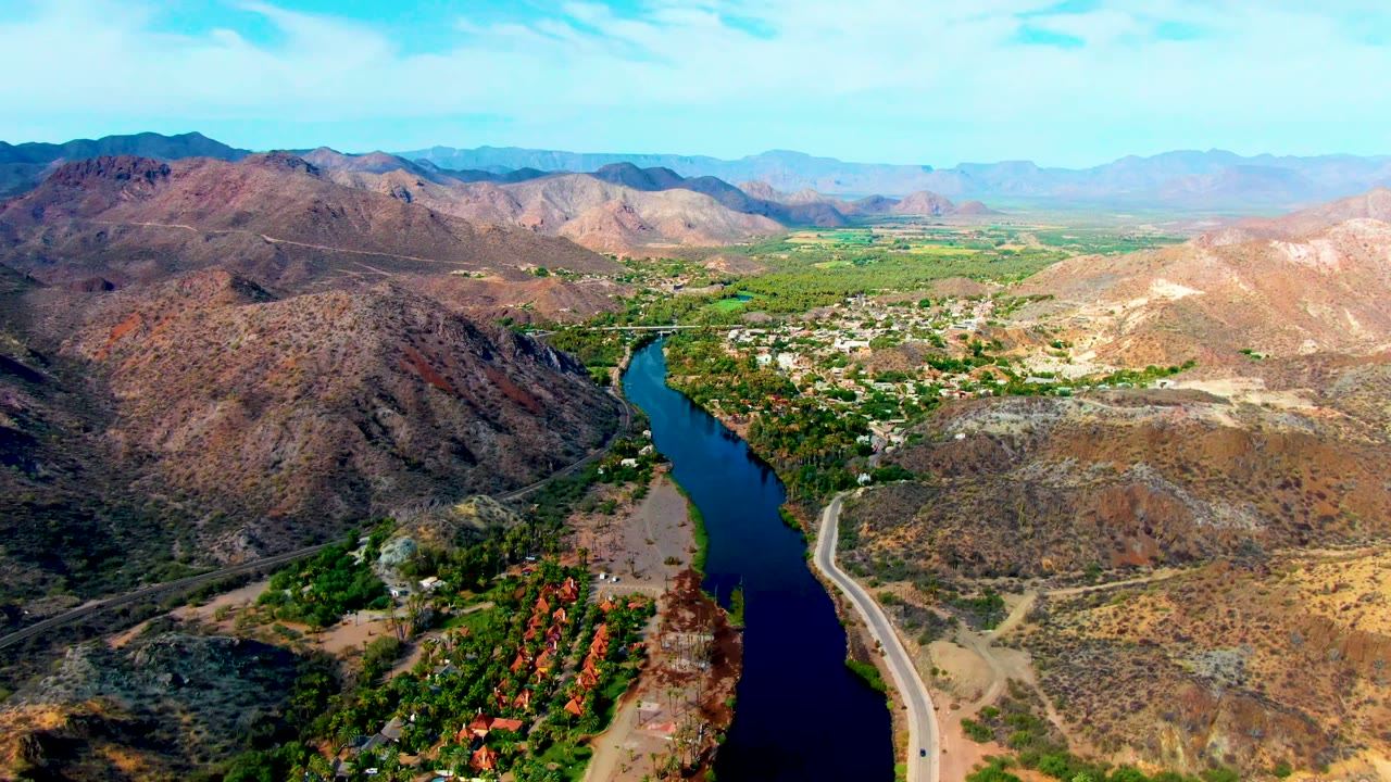 Opening aerial frame over Mulege, Baja California Sur, with a dark blue river running beside a road through dry rocky hills and no visible people.