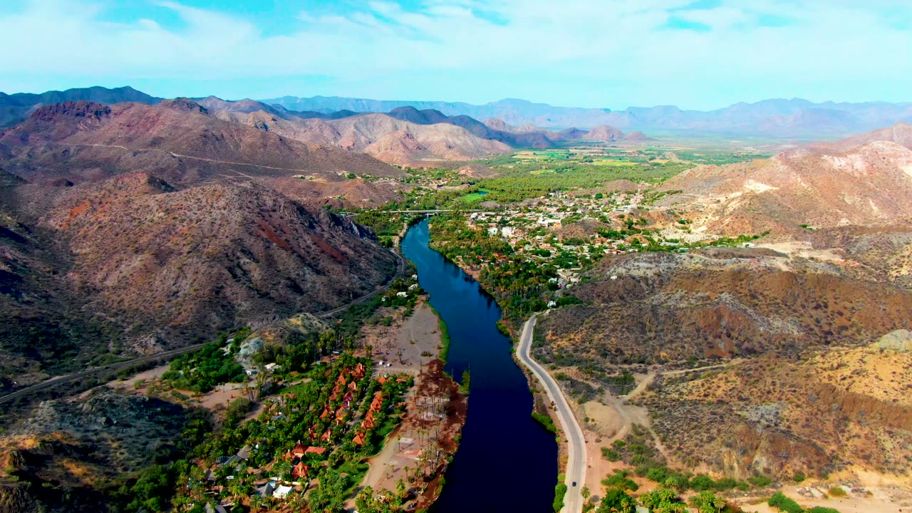 Mid-clip aerial frame over Mulege with the river centered between brown hills, a pale road along the water, green valley patches, and no visible people.