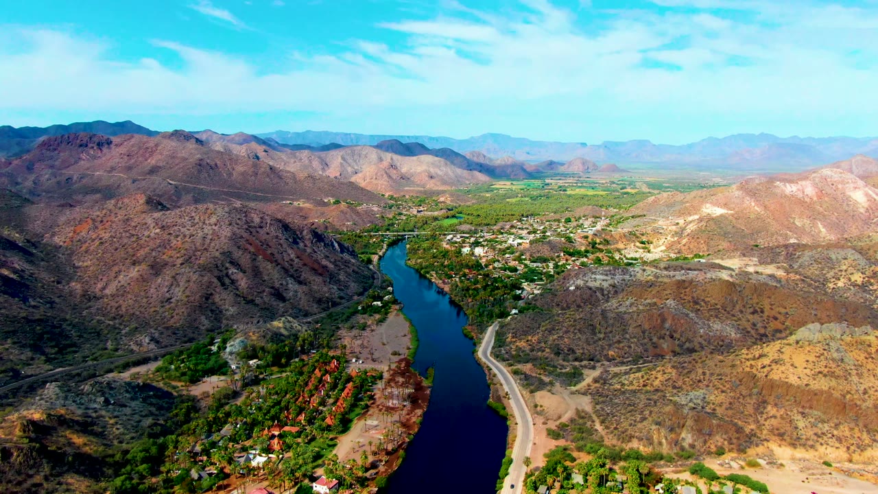 Opening aerial frame over Mulege, Baja California Sur, showing a blue river bending between rocky brown hills, a roadside along the bank, patches of green vegetation, scattered structures, and no visible people.