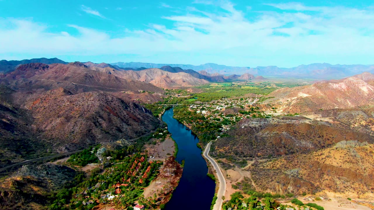 Wide aerial landscape with river, road, green valley, and layered mountains.