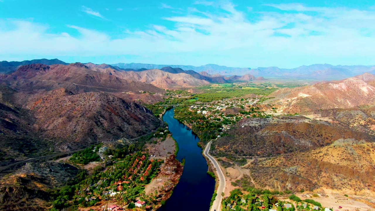 Mulege, Baja California Sur aerial frame with the dark blue river centered below the hills, green trees and small structures along the water, a pale road on one bank, dry ridges beyond, and no visible people.