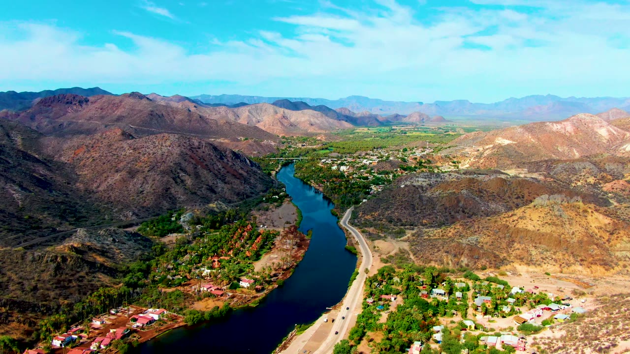 Aerial view over Mulege, Baja California Sur, showing a dark river curve, parallel highway, rocky hills, scattered buildings, green vegetation, and no visible people.