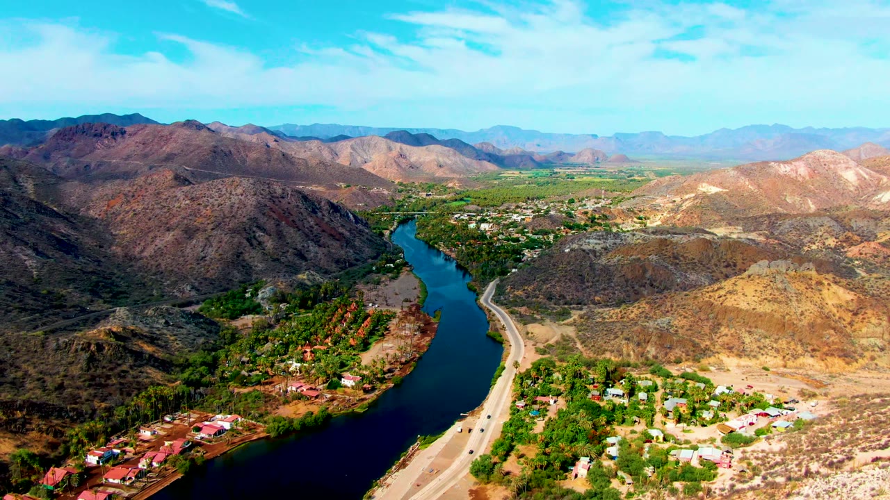 Wide aerial valley view with river, highway, vehicles, hills, and scattered buildings.