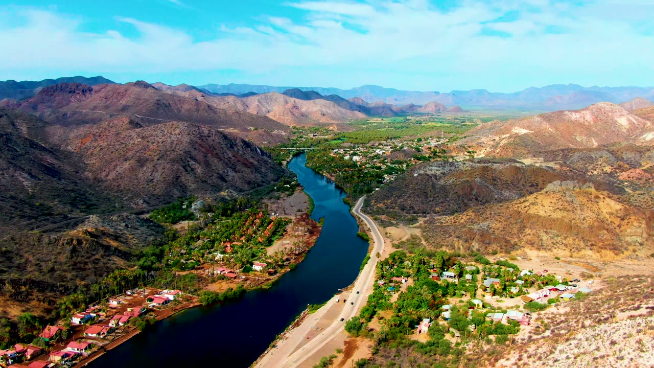 Elevated Mulege view with the river bending through the center, a pale highway and vehicles along the bank, green settlement areas near the water, and no visible people.
