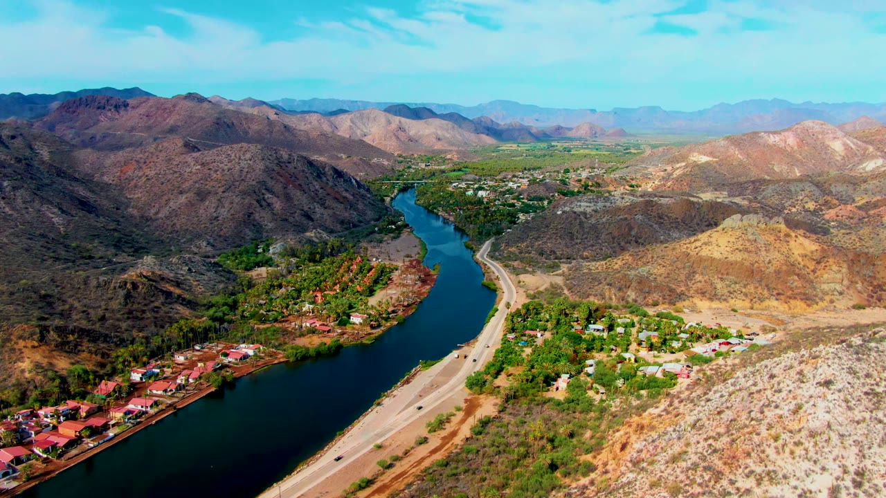 Opening aerial frame over Mulege, Baja California Sur, showing a blue-green river beside a paved road, riverside houses, dry mountain slopes, distant hills, and no visible people.