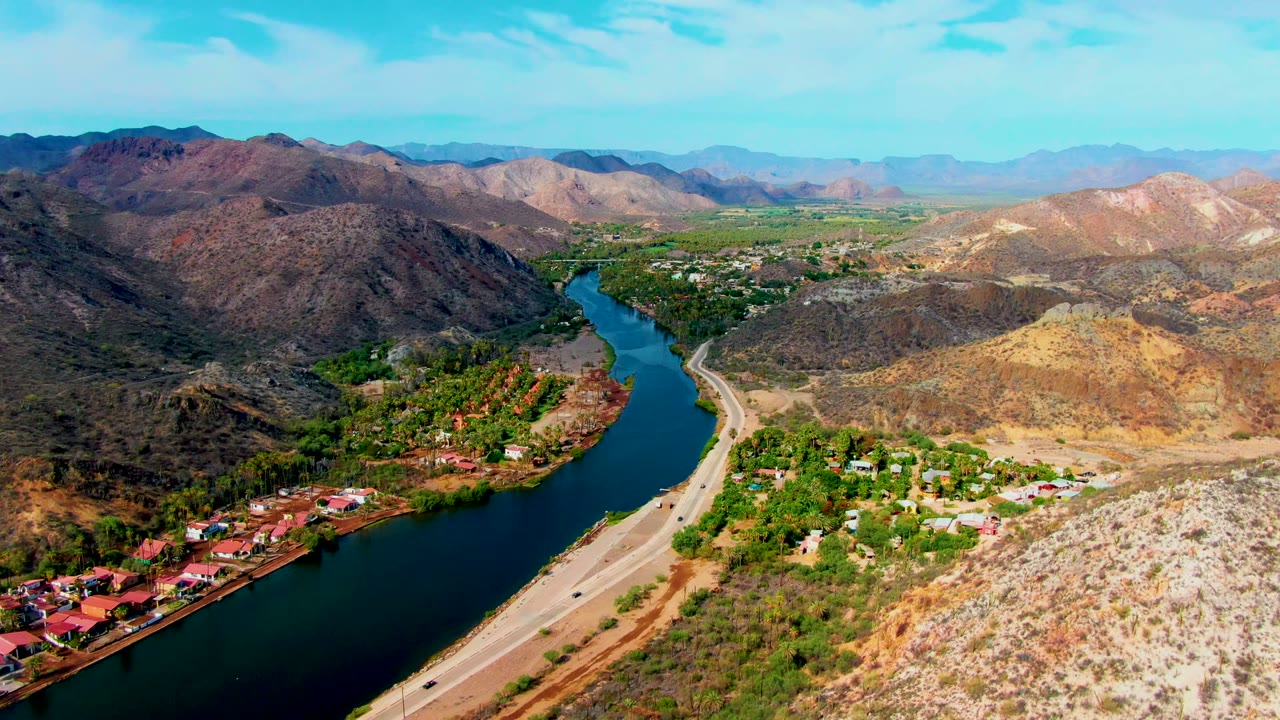Mulege aerial frame showing small cars on the road beside the dark river, red-roofed houses along the bank, green trees, tan desert slopes, mountains, and no visible people.