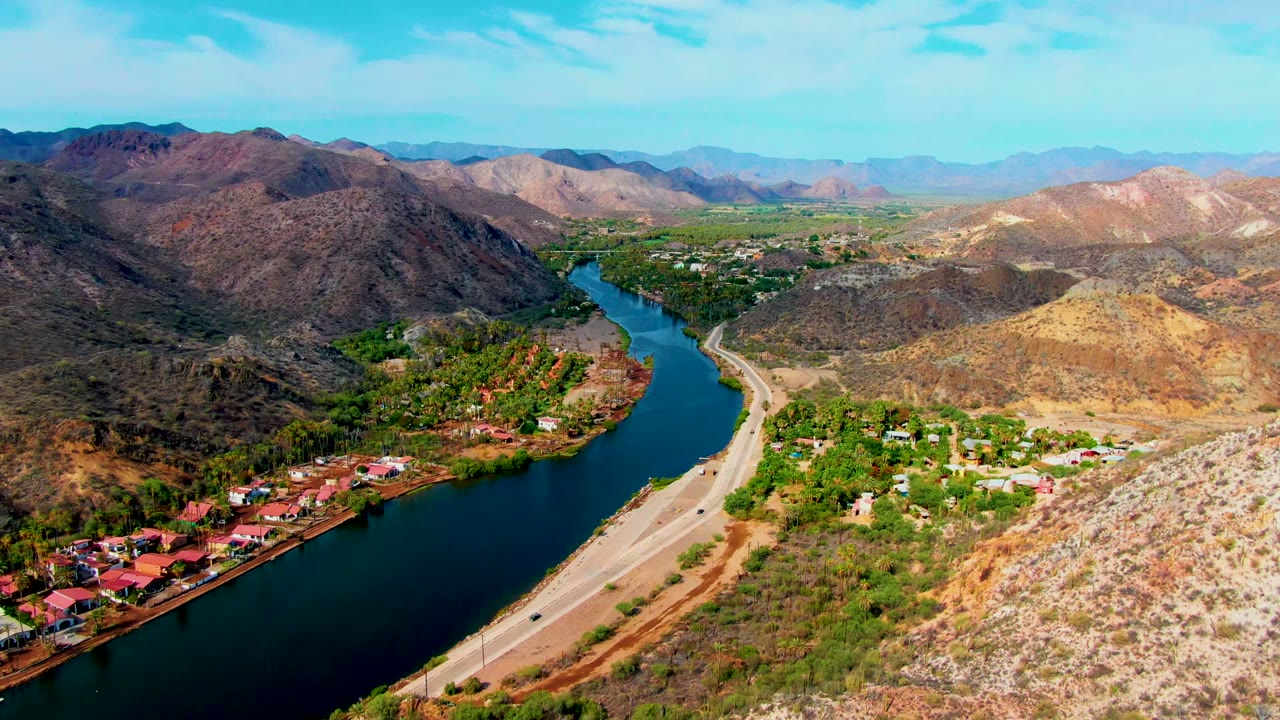 Aerial view over Mulege, Baja California Sur, showing a blue river bending past a road, green vegetation, red-roofed buildings, dry hills, and distant mountains with no visible people.