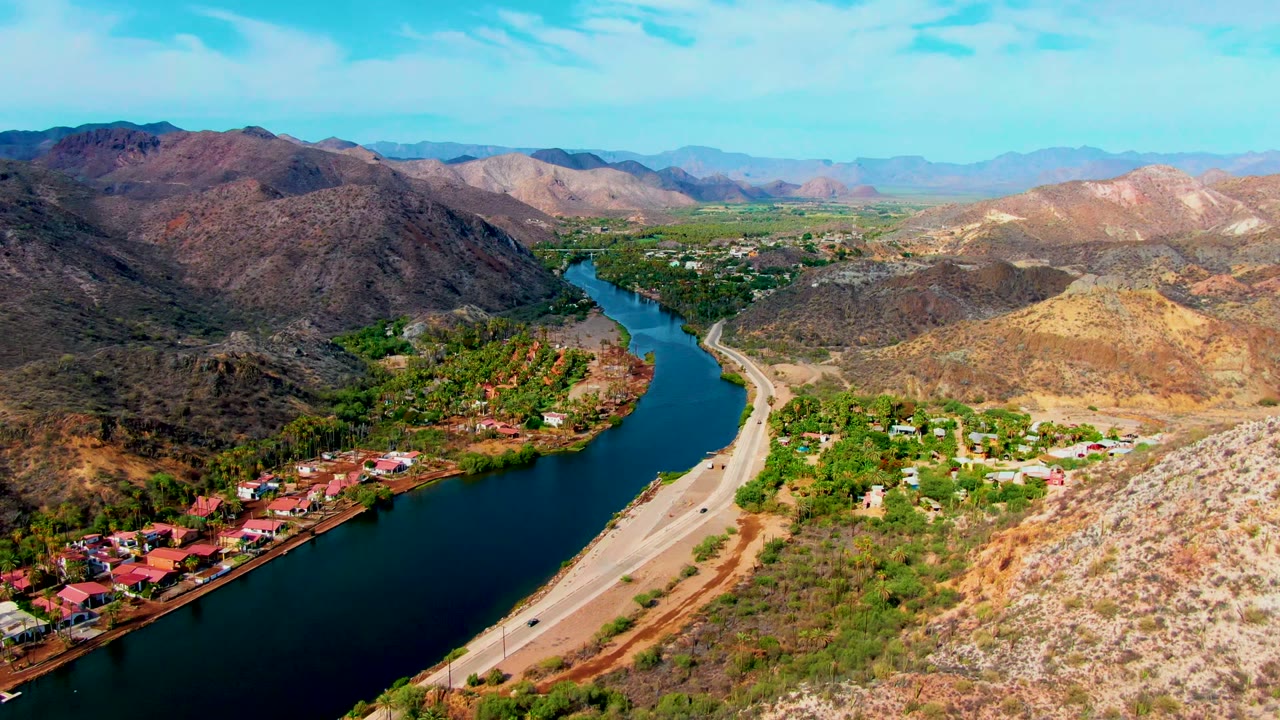Wide aerial river bend beside a road with houses and mountain ridges.
