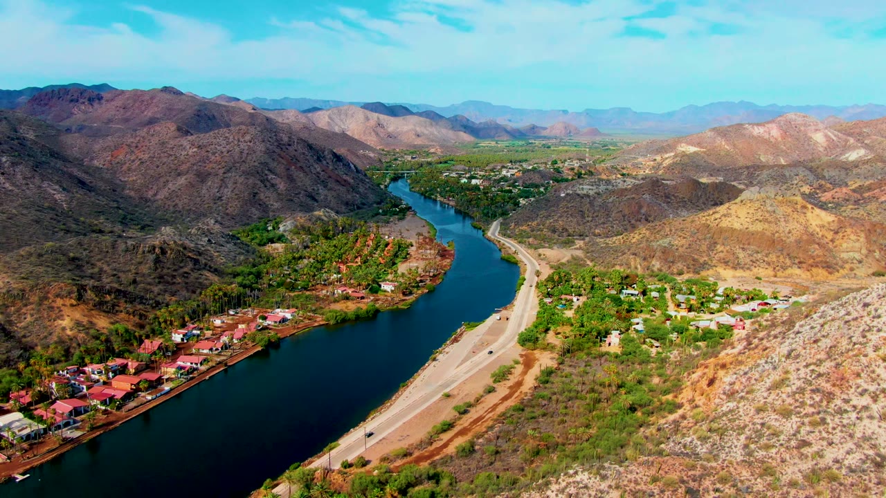 Drone-height view over Mulege with dark blue water in the foreground, a road curving beside it, clustered riverside buildings, tan hills, distant mountains, and no visible people.