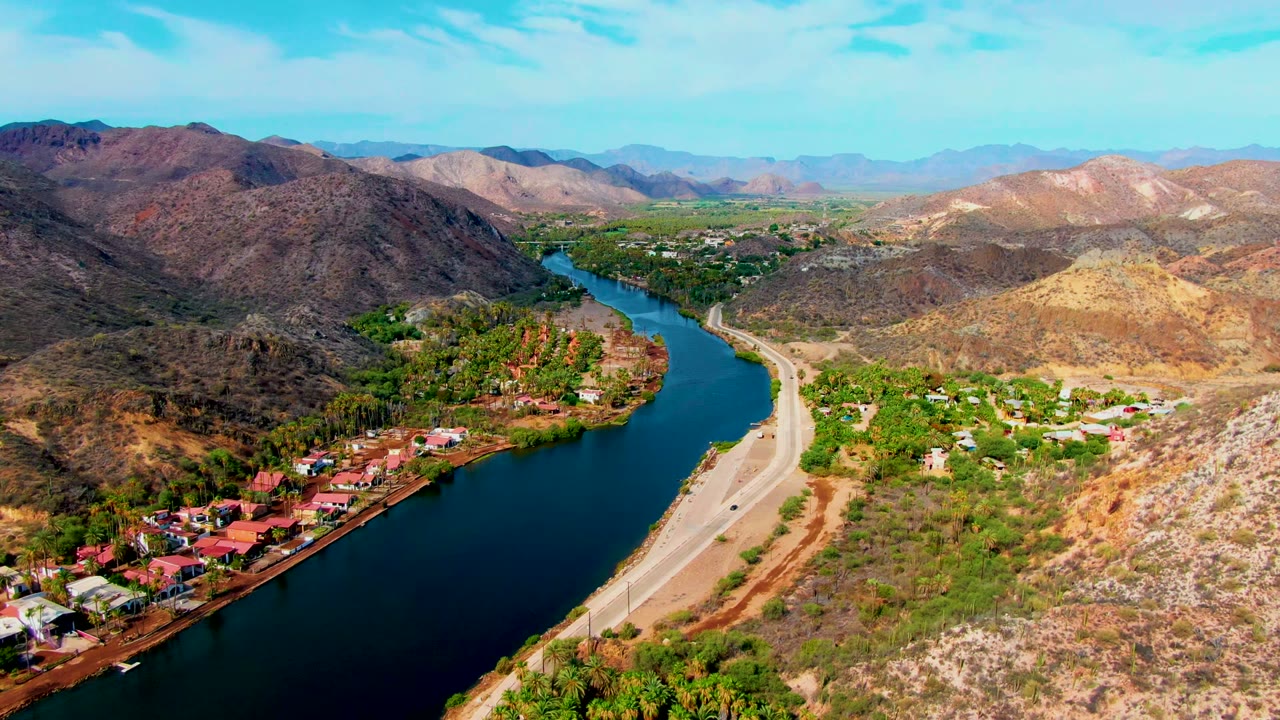 Wide aerial view in Mulege, Baja California Sur, with a blue river curving past a roadside neighborhood, dry hills, distant mountains, and no visible people.