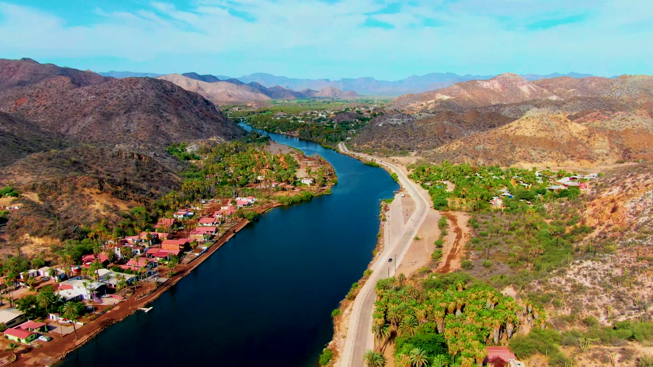 Aerial view over Mulege, Baja California Sur, with a dark blue river beside a pale road, small homes, palms, dry hills, and no visible people.