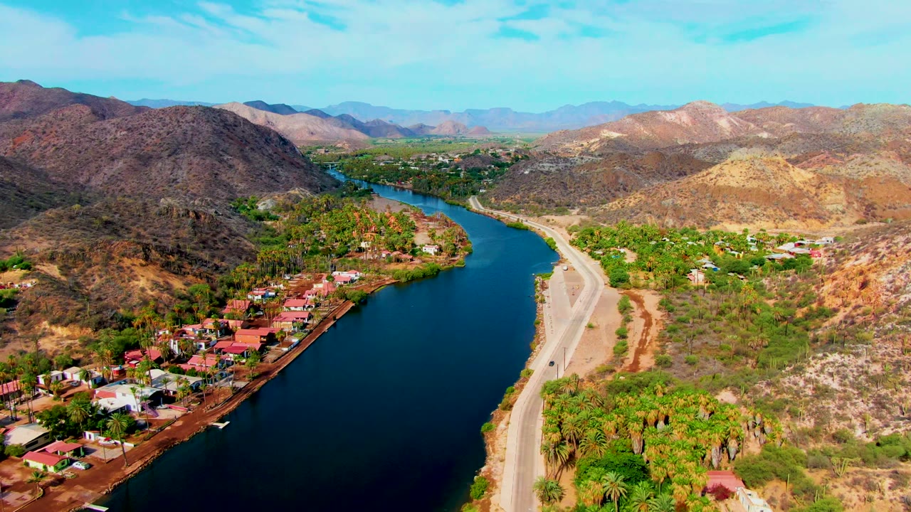 Wide aerial view of river bend, roadside, homes, palms, and arid hills.