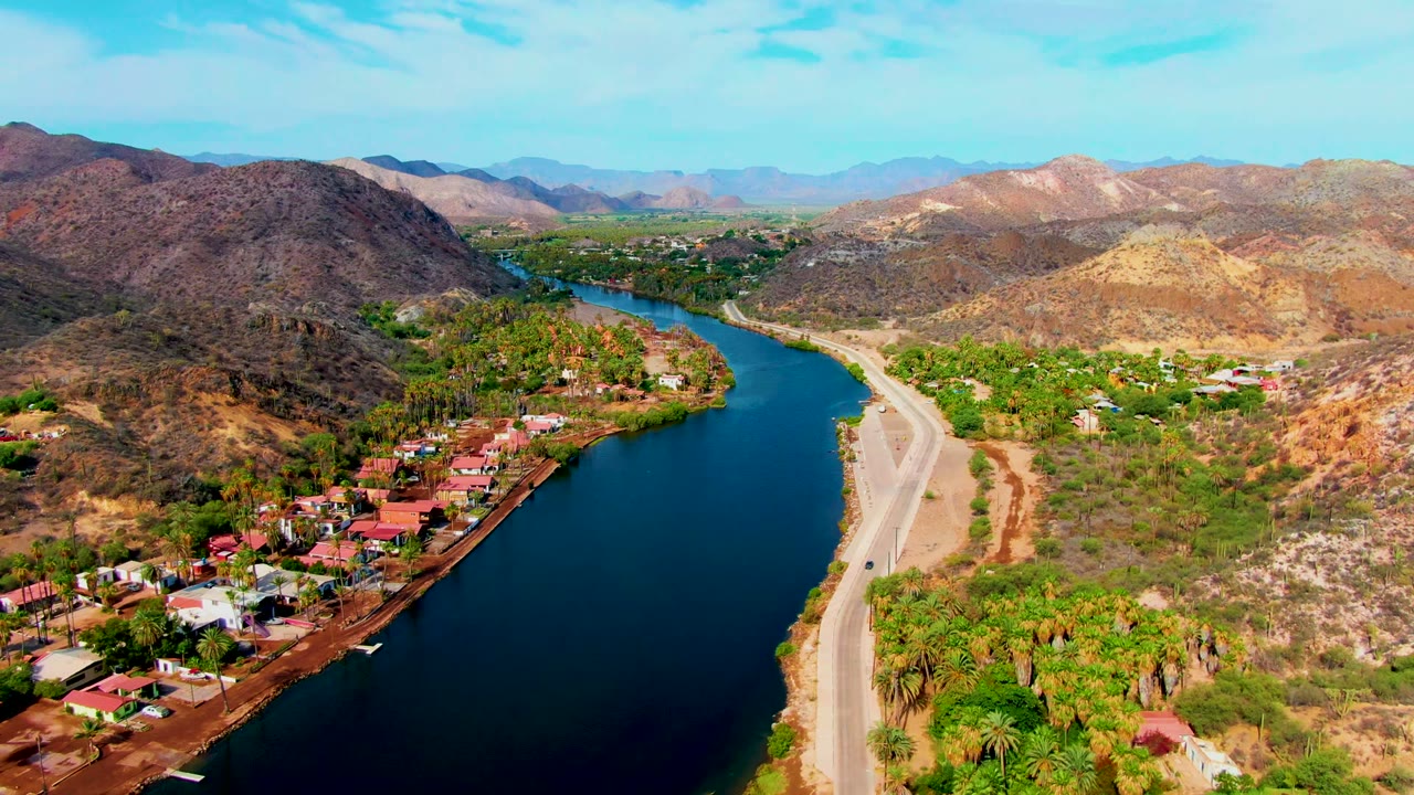 Drone view over Mulege, Baja California Sur, with dark river water in the lower frame, a road on the bank, village homes, green palms, dry hills, and no visible people.