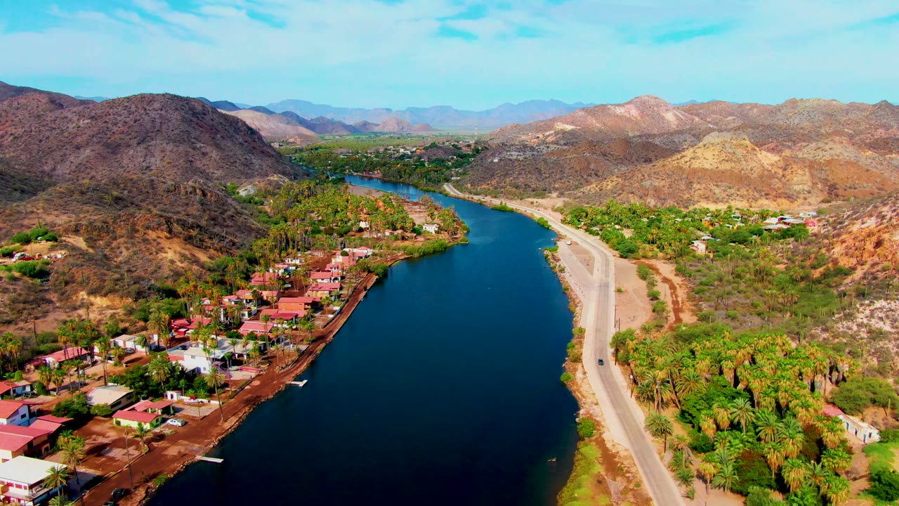 Aerial view over Mulege, Baja California Sur, with a dark blue canal at left, a pale road at right, red-roofed homes and palms below, dry hills beyond, and no visible people.