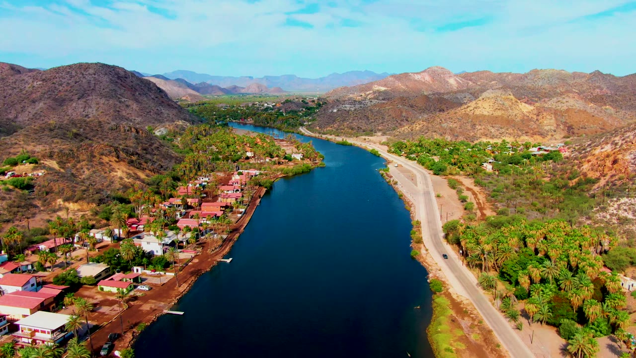 Aerial Mulege frame with the blue canal filling the lower left, roadside greenery and palms in the center, homes along the bank, a pale road on the right, and no visible people.