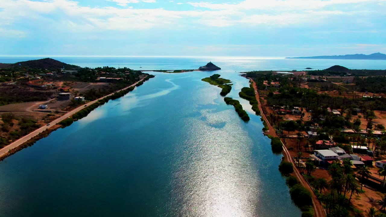 Opening aerial frame over a blue canal in Mulege, Baja California Sur, with roads on both banks, low buildings among palms, hills and distant water on the horizon, and no visible people.