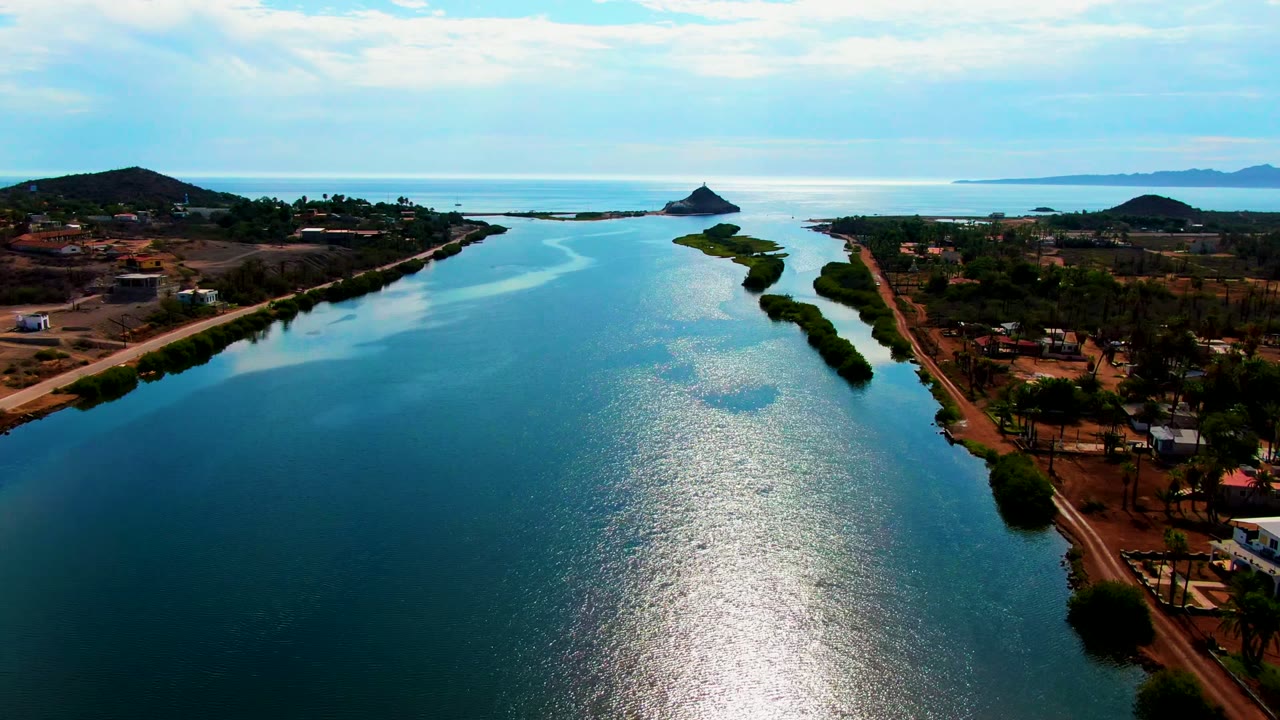 Opening aerial frame over a blue waterway in Mulege, Baja California Sur, with tree-lined roads on both banks, low hills, distant coast, bright water reflection, and no visible people.