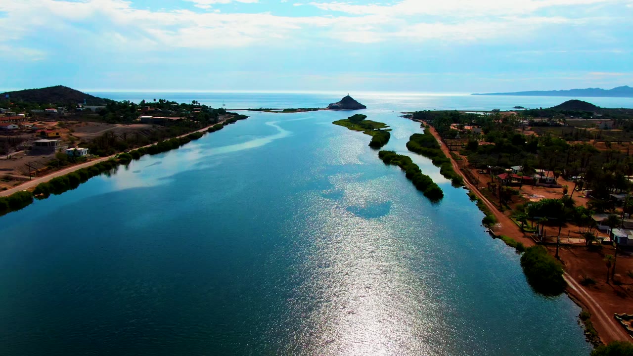 Wide aerial frame over the Mulege waterway with the channel centered, rows of trees and roads along the edges, scattered structures on the right bank, distant islands or coastal landforms, and no visible people.