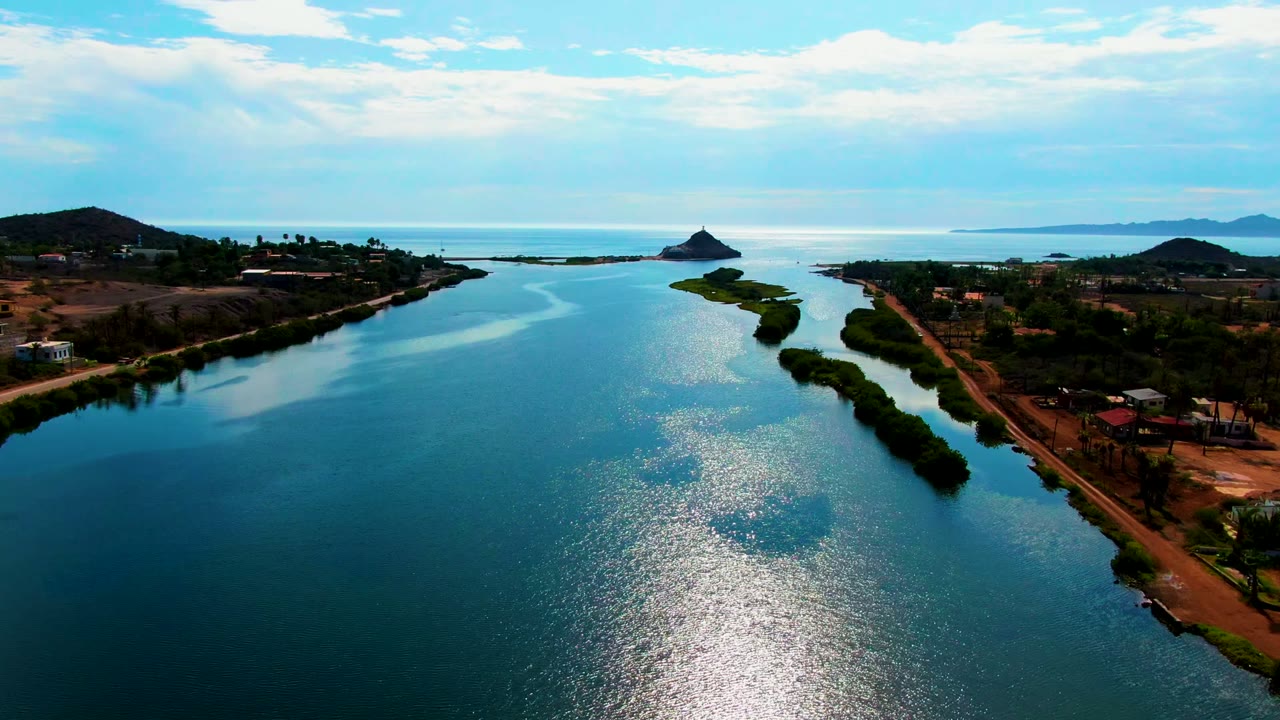 Opening aerial frame over a calm Mulege, Baja California Sur water channel with bright reflection on the water, a narrow green island, shoreline buildings, low hills, and no visible people.