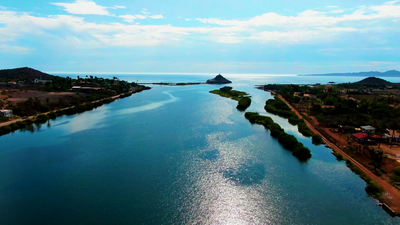 Calm waterway with a vegetated island and shoreline buildings under a cloudy sky.
