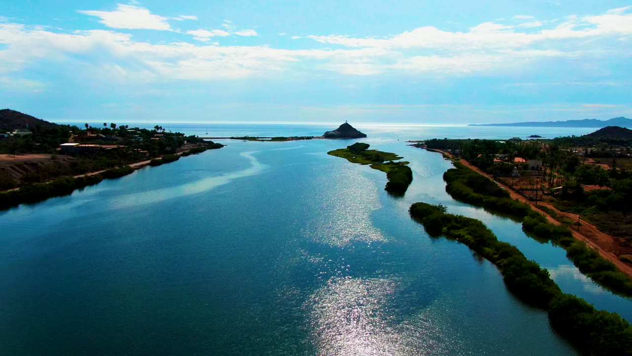 Aerial view in Mulege, Baja California Sur, with blue river water in the foreground, a narrow green island through the center, coastal land on both sides, distant hills, clouds, and no visible people.