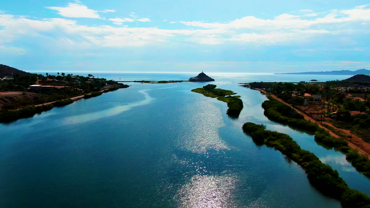 Wide aerial river view with sunlit water, green island, shoreline roads, and distant coast.